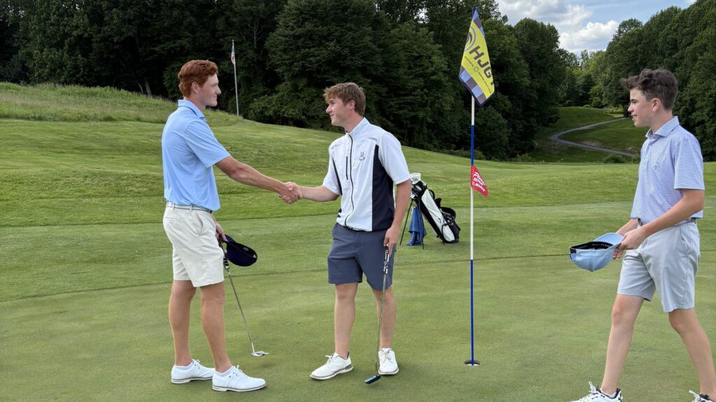 Trzej młodzi gracze na polu golfowym, dwóch ściska sobie dłonie przy dołku z flagą po skończonej grze