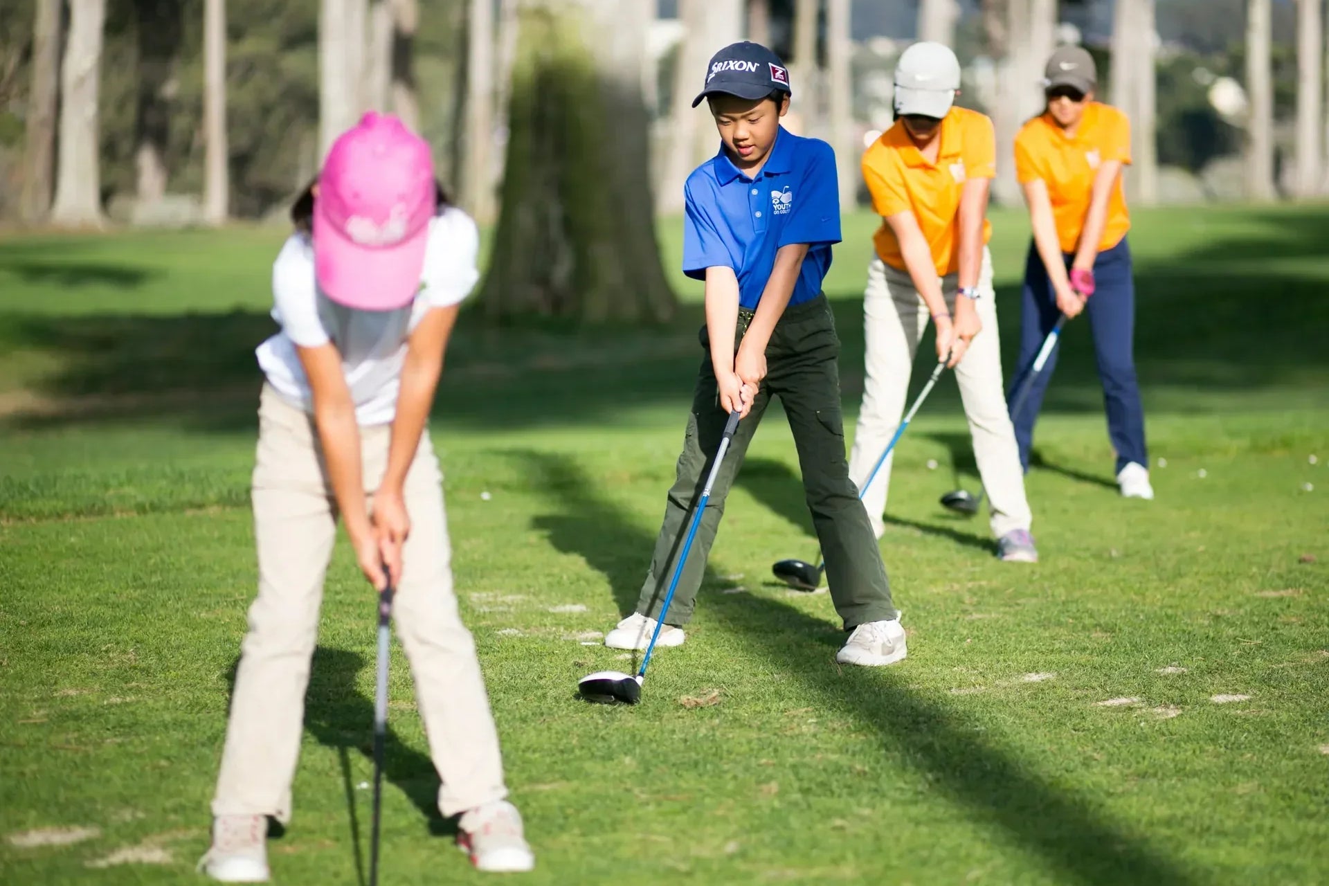 Grupa dzieci ćwiczących uderzenia golfowe na trawiastym polu golfowym podczas zajęć w szkółce