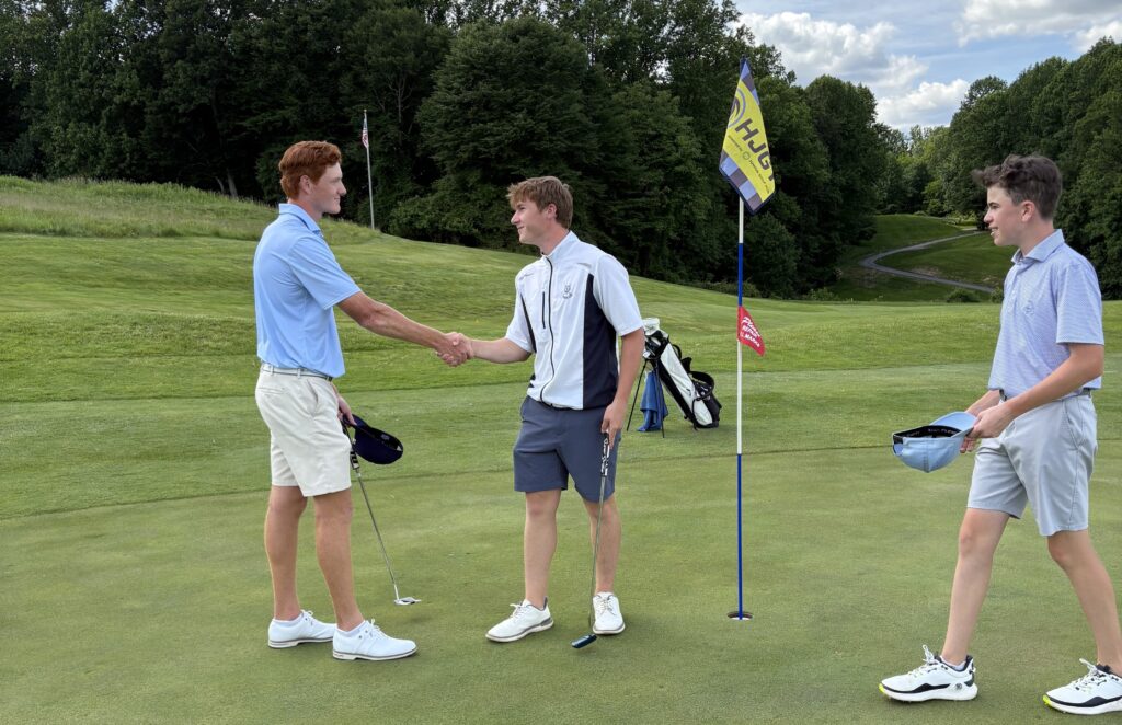 Trzej młodzi gracze na polu golfowym, dwóch ściska sobie dłonie przy dołku z flagą po skończonej grze