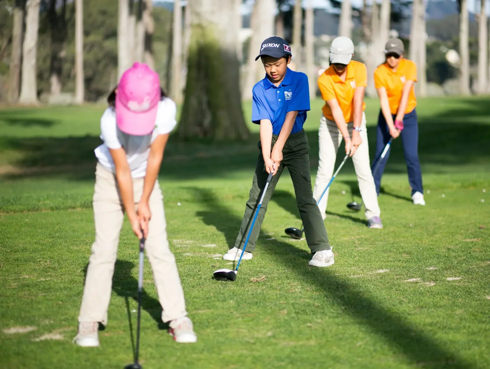 Grupa dzieci ćwiczących uderzenia golfowe na trawiastym polu golfowym podczas zajęć w szkółce